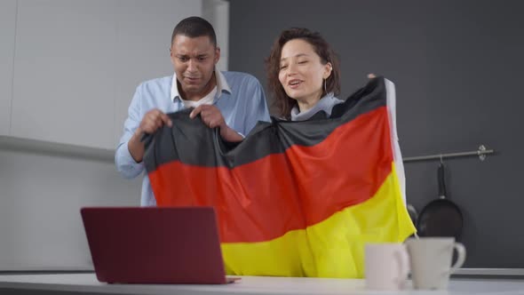 Joyful Interracial Couple with German Flag Cheering for Soccer Team Watching Online Match on Laptop alt