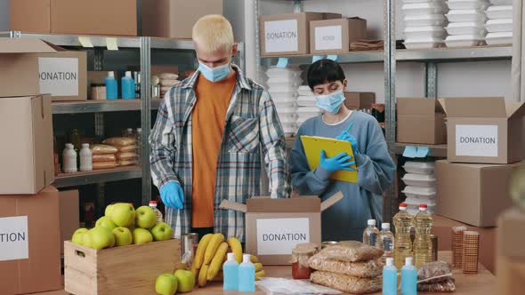 Man Packing Donation Box While Woman Using Clipboard alt