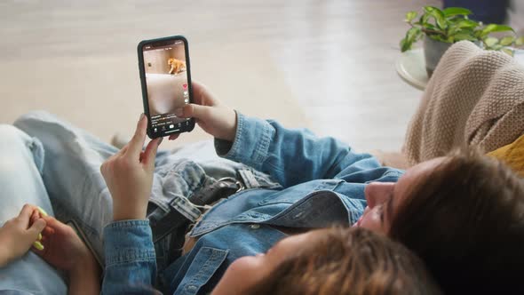 Rearview of Young Women Sitting on Sofa and Watching Short Funny Videos on Mobile Phone alt