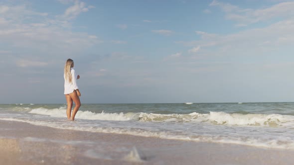 A Slender Girl in a Gentle Blue Swimsuit and Shirt Walks Along the Sandy Beach Near the Blue Sea