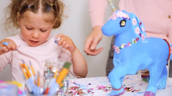 Step by step. Mother and daughter decorating paper mache unicorn with jewels and paper flowers alt