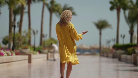 a Curly Blonde in Sunglasses and a Yellow Fluttering Dress Looks at the Phone and Walks Away Against alt