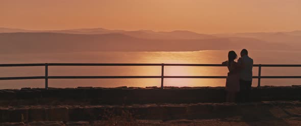 Young couple watching sunset above the lake from the viewing point. Wide shot alt