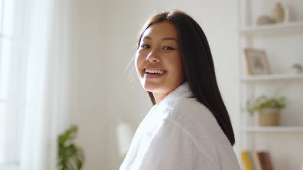 Young Peaceful Korean Woman Wearing White Bathrobe Smiling to Camera Posing at Home in Morning alt