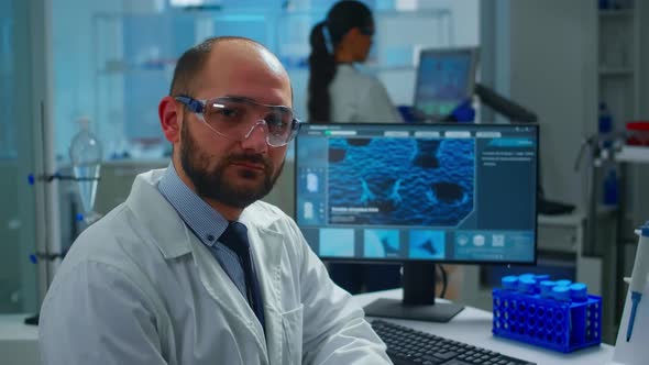 Portrait of Exhausted Scientist Man Looking at Camera Sitting in Front of Computer alt