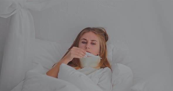 Young Woman Lies in Bed with a Scarf and a Thermometer is Sick