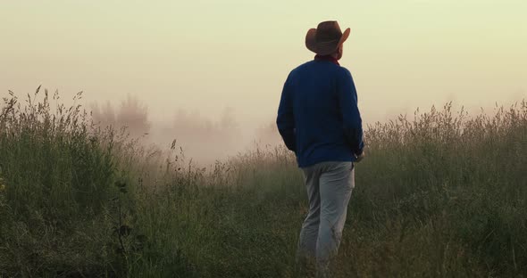 Man in a Cowboy Hat Stands in a Foggy Field at Sunset and Lights a Cigarette
