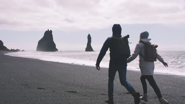 Young Couple Holding Hands and Running on Black Sand Beach in Iceland Slow Motion alt