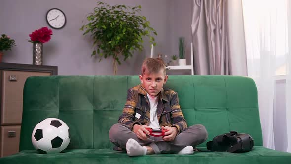 Modern Teen Boy Sitting on Comfortable Sofa at Home and Applying Joystick while Playing Video Game alt
