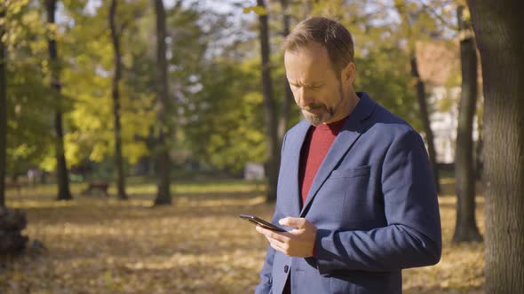 A Middleaged Handsome Caucasian Man Works on a Smartphone in a Park in Fall