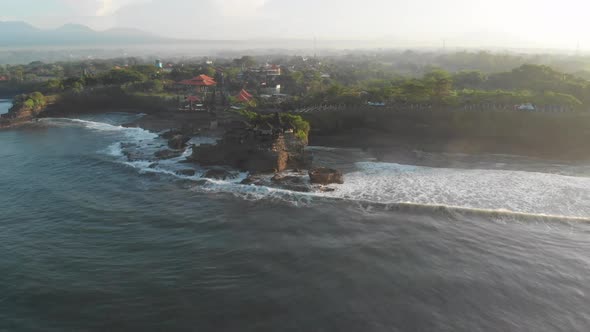 Aerial pan view of waves crashing against a temple with mountain views in the distance