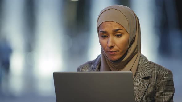 Portrait of Muslim Business Woman User in Hijab Sits in Evening City Outdoors Working with Laptop alt