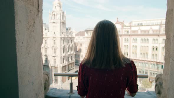 Young Woman Walks on Hotel Balcony Looks at City Street and Smiles to Camera