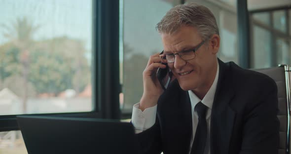 Businessman in formal wear and glasses talking on the phone, smiling,  while working at the office. alt