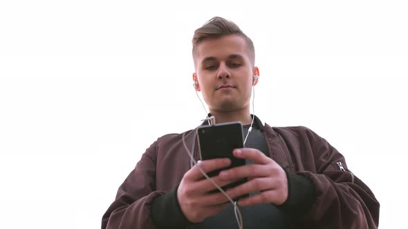 Young Handsome Man Holds Mobile Phone Listens to Music on Beach During Sunset