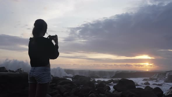 Girl in Denim Shorts Stands and Takes a Photo on Her Phone of a Huge Wave Crashing Against Rocks alt