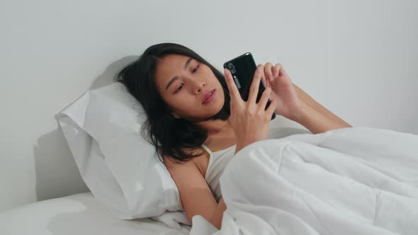 Young Asian woman using smartphone checking social media feeling happy smiling while lying on bed. alt