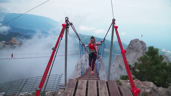Young Athletic Girl Is a Traveler Crossing the Rope Bridge Above the Precipice in the Mountains alt