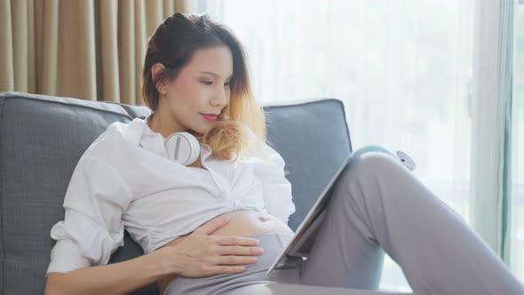 Asian young woman pregnant sitting and reading book with smile on sofa. alt