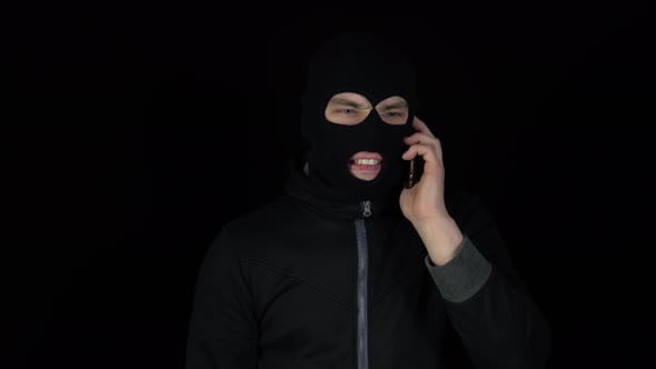 A Man in a Balaclava Mask Is Talking on a Phone. A Thug Screaming on the Phone on a Black Background alt