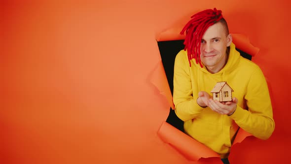 Young Handsome Man with Small Wooden House Sticking Out of Hole of Orange Background alt