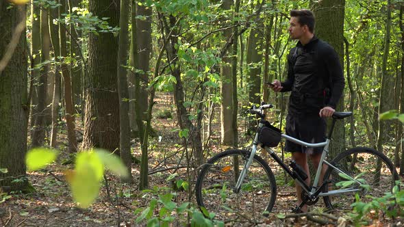 A Cyclist Stands with a Bike in a Forest and Works on a Smartphone alt