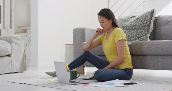 Focused asian woman sitting on floor and working remotely from home with smartphone and laptop alt