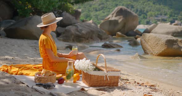 Pretty Woman in Hat and Yellow Dress Using Her Phone Having Picnic Summer Day on Lonely Beach alt