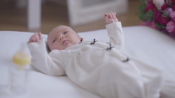 Relaxed Caucasian Little Girl in White Suit Lying in Bed with Bouquet of Flowers and Baby Bottle alt