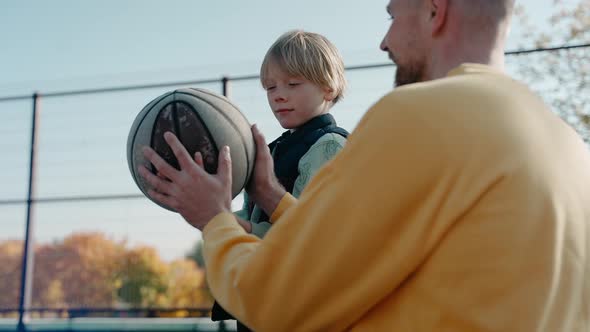 Father Explaining to Son How to Hold and Throw a Basketball Ball on a Sunny Day