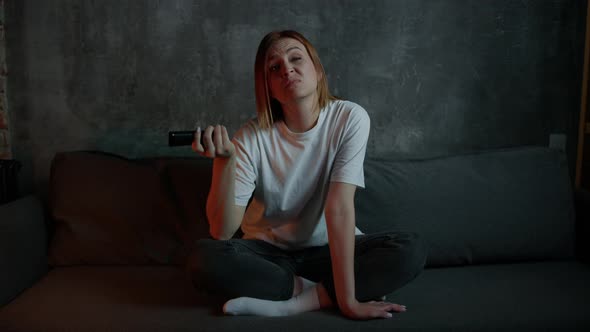 A Young Woman is Sitting on the Couch and Can't Choose What to Watch on TV alt