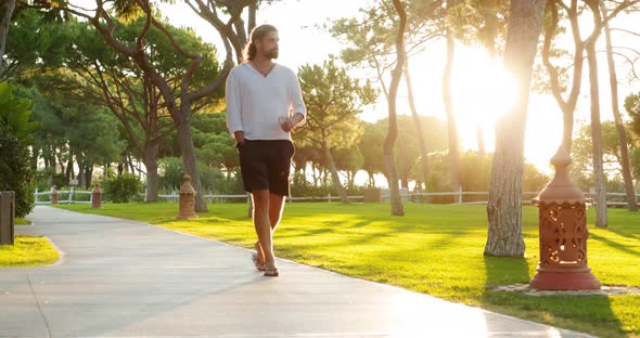 Caucasian Guy Wearing Casual White Shirt and Blue Shorts Walks Along the Park alt