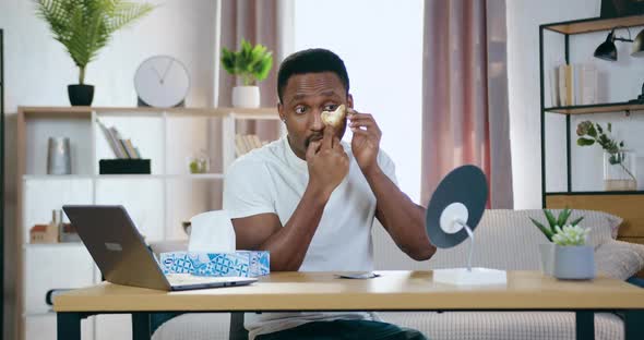 African American Sitting in front of Small Mirror at Home and Putting Golden Patchi Under eyes alt