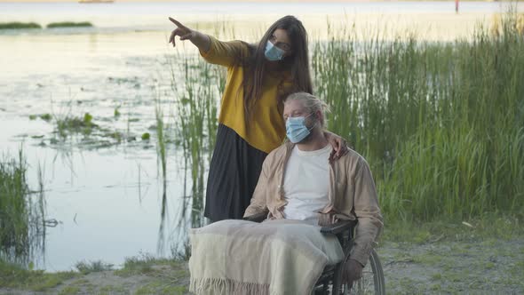 Portrait of Confident Young Woman in Face Mask Talking with Paralyzed Man in Wheelchair. Wide Shot alt