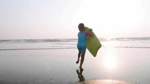 Little Girl is Running on the Sea to Waves Holding Surfboard in Her Hands