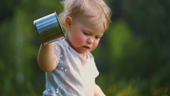 one year old baby spills liquid from a metal mug. The concept of walking outdoors, alt