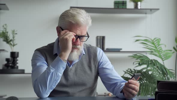 An Elderly Man with a Gray Beard Uses a Smartphone in the Office alt