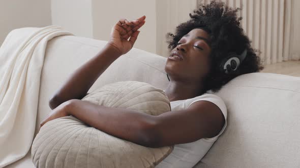 Calm Happy Young Black Race Woman in Headphones Sitting on Comfortable Sofa with Eyes Closed alt