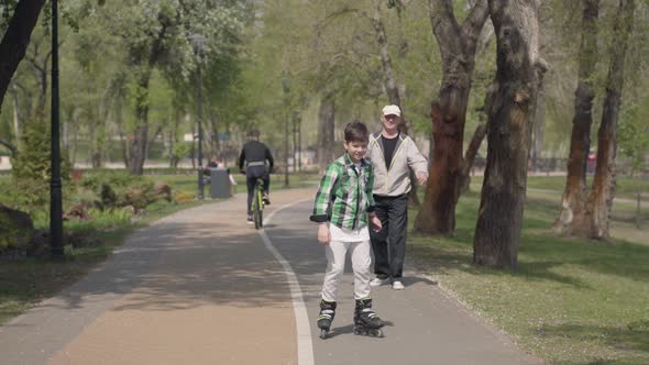 Cute Boy in Bright Clothes Rollerblading in the Park, His Grandfather Walking Near alt