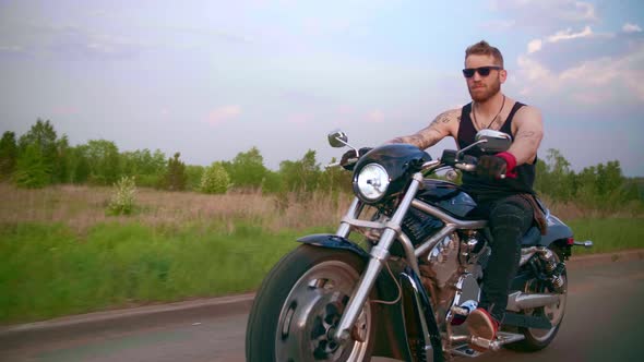 Stylish Biker with Tattoos Rides a Motorcycle on a Country Road at Sunset