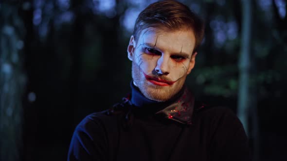 Spooky Scary Man with Vampire Face Paint Standing in Night Forest Looking at Camera on Halloween alt