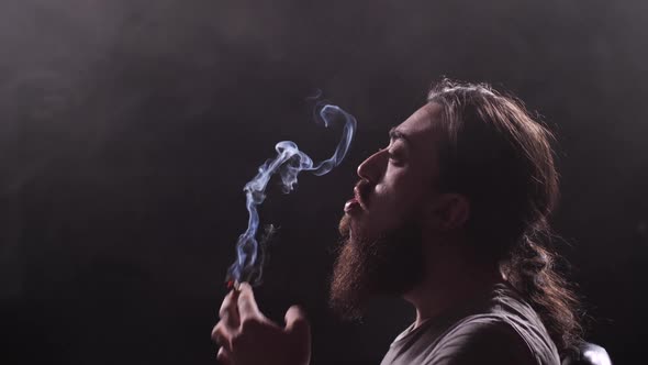 A Guy with a Long Beard and Oblique Smokes a Cigarette on a Black Background alt