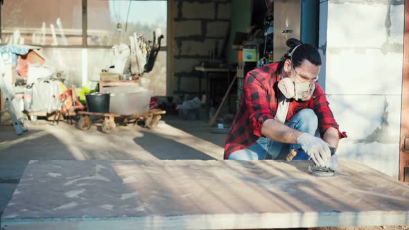 A Young Man in a Construction Respirator Grinds a Parquet Board with a Sander alt