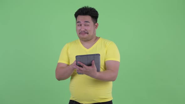 Happy Young Overweight Asian Man Thinking While Using Digital Tablet alt