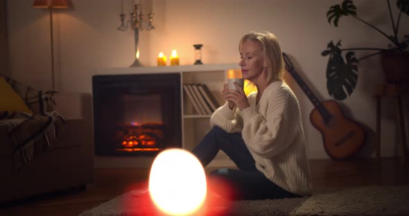 Smiling Aged Woman Drinking Cup of Hot Coffee Sitting in Front of Fireplace alt