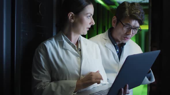 Diverse female and male it technicians in lab coats using laptop checking computer server alt