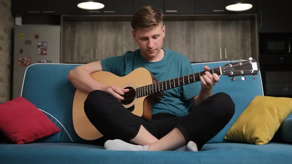 Caucasian Man Sitting on a Blue Sofa and Playing at the Guitar. alt