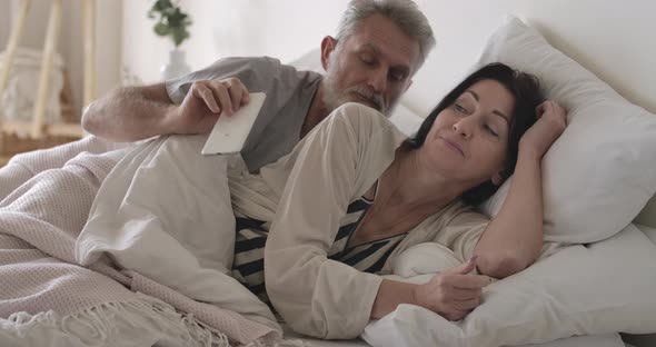 Furious Caucasian Senior Man Taking Away Wife's Smartphone in Bed. Husband Looking Angrily at alt