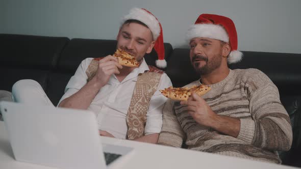Happy Gay Male Couple with Santa Hats Watching Movies on the Laptop and Eating Pizza alt