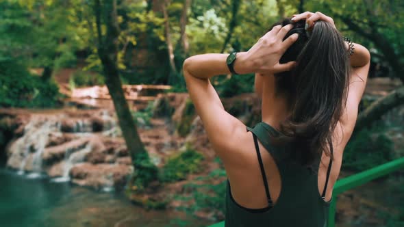 Fit Woman Fixing Her Hair While Enjoying Sunny Day in the Forest, Watching Forest Waterfalls alt